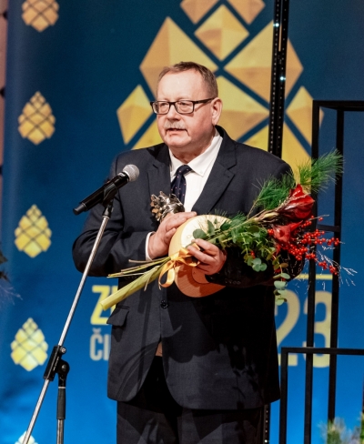 Vīrietis ar ziediem un balvu rokās runā mikrofona